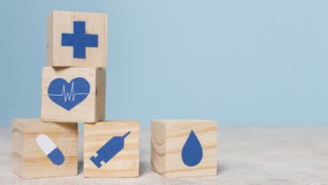 pięć drewnianych klocków nadrewnianym blacie, klocki mają niebieskie grafiki związane z medycyną (serce, tabletka, strzykawka itp.). W tle niebieska ściana 