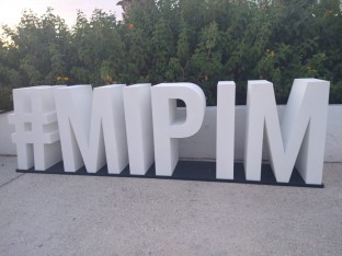 białe litery tworzące napis #MIPIM ustawoione na betonowej płycie, w tle zieleń drzew 